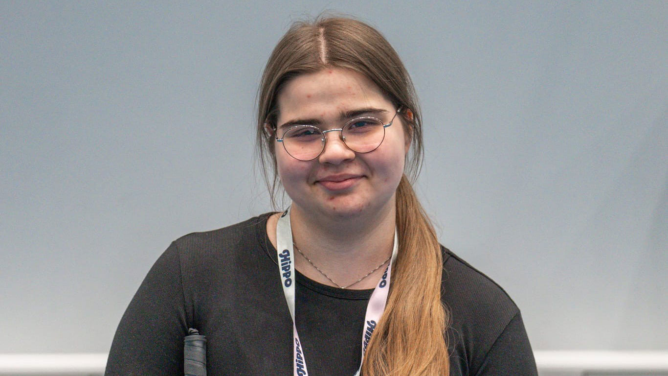 Silmälasipäinen tyttö pitkät hiukset poninhännällä hymyilee kisapaikalla lähikuvassa.